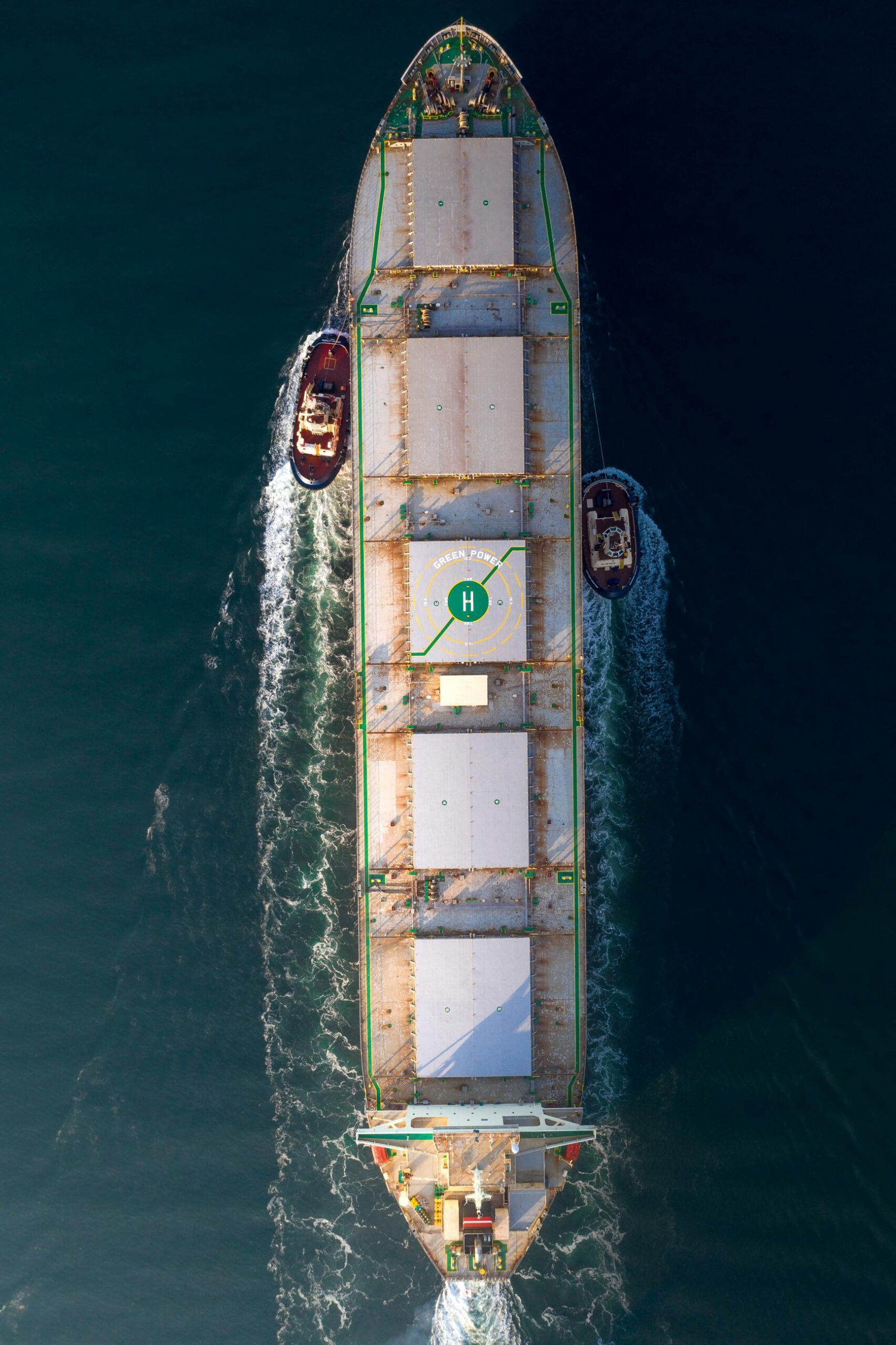 An aerial photo of a large cargo ship with tugboats assisting in open sea, showing maritime transportation.
