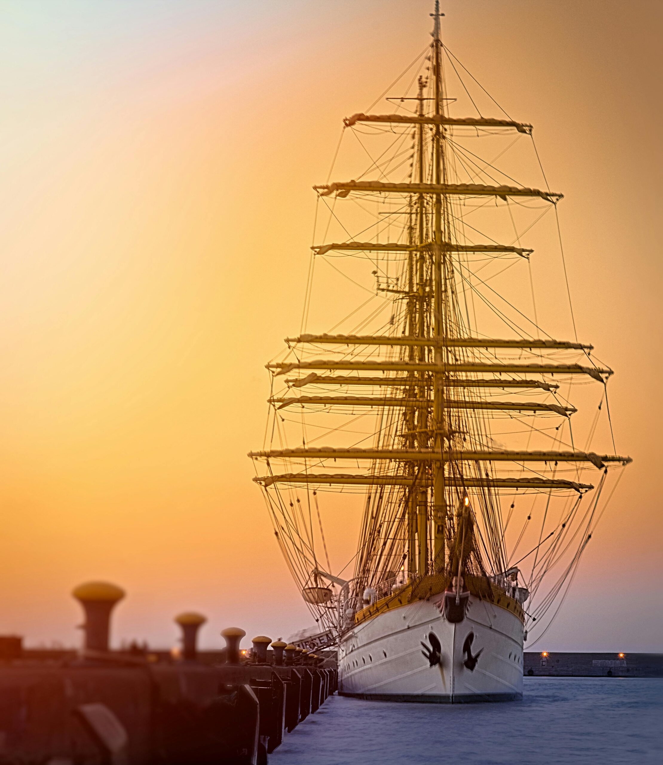 Home A full-rigged ship anchored at sunset in a tranquil harbor, capturing nautical elegance.