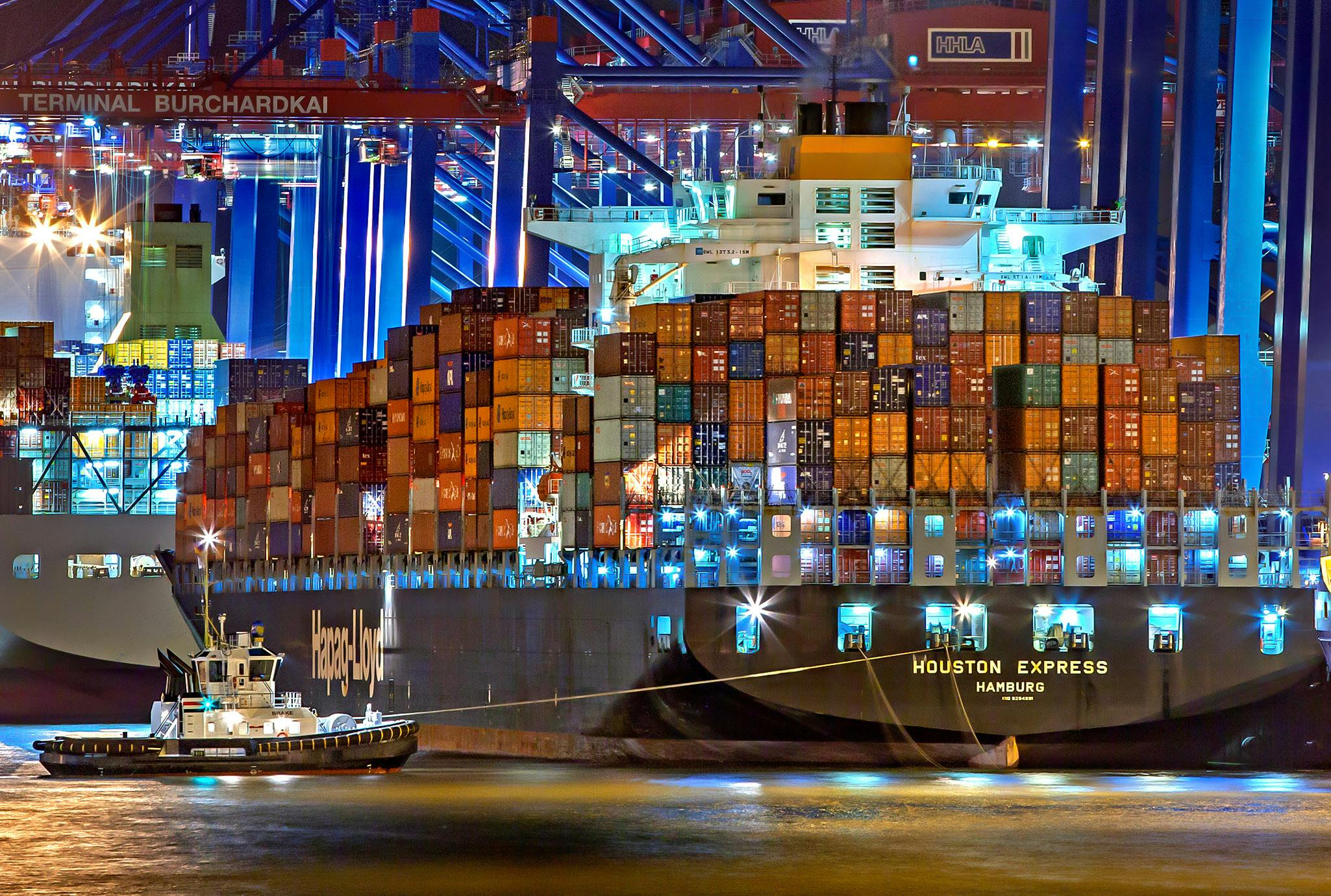 Home A brightly lit cargo ship at Hamburg harbor with stacked containers and a tugboat.