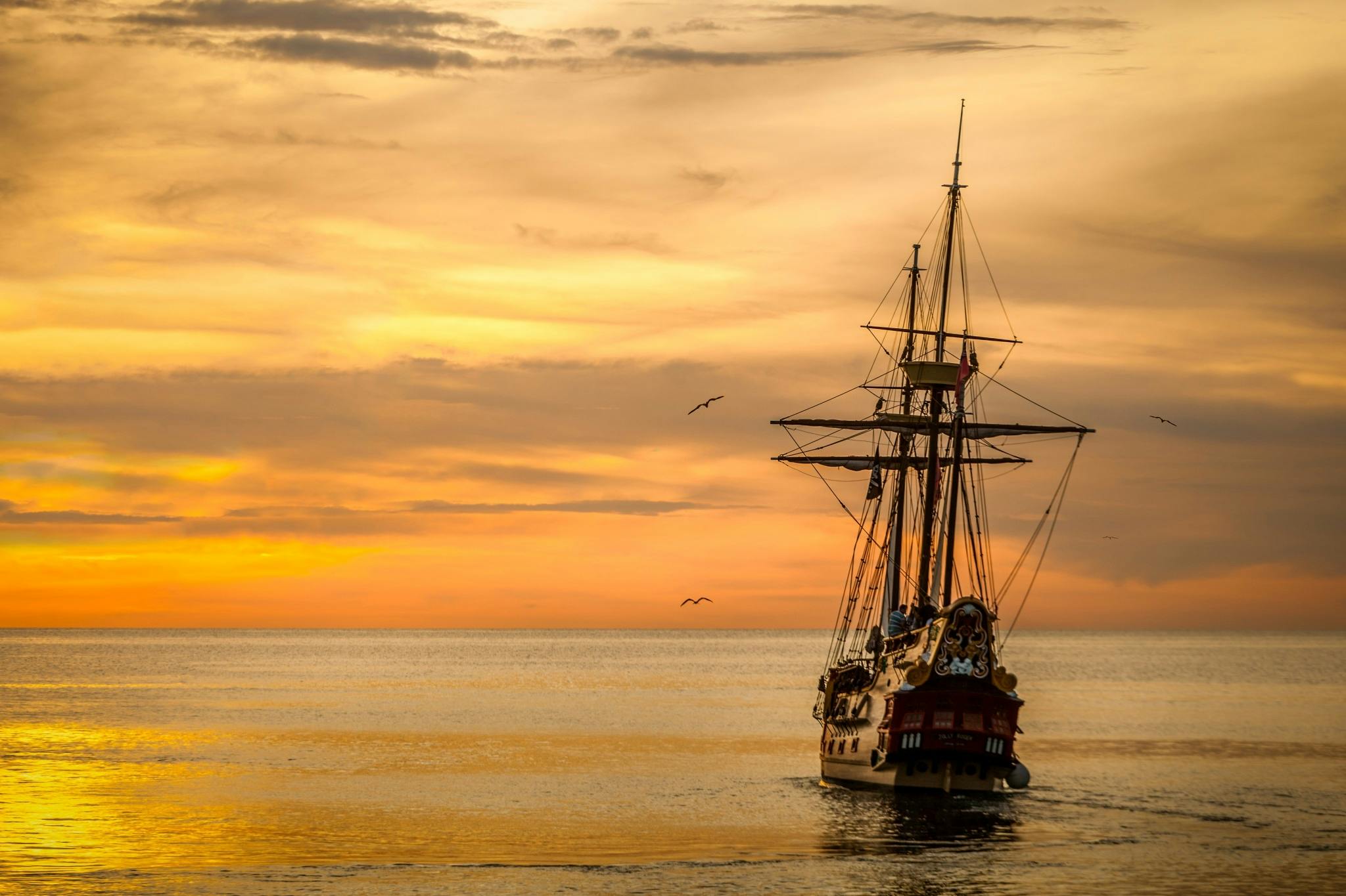 Home A majestic pirate ship sails into the serene waters at sunset, casting a dramatic silhouette.